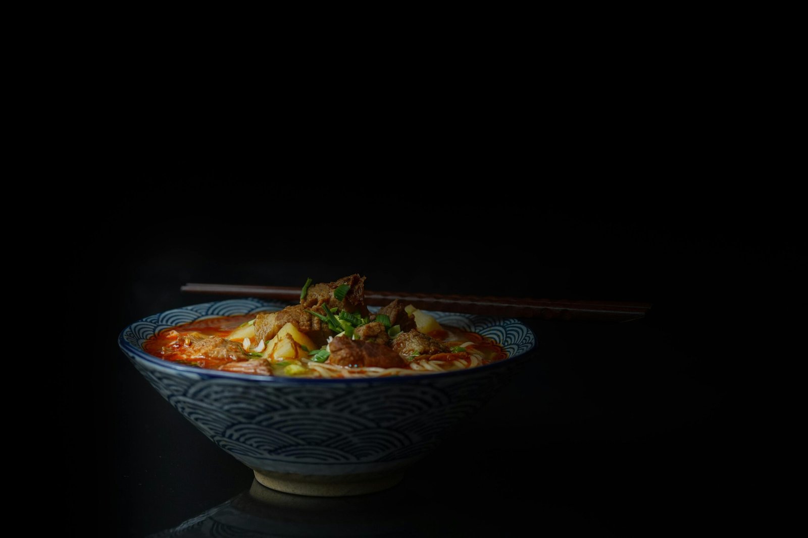Close-up of a flavorful Asian noodle soup with meat and vegetables, served in a patterned bowl on a dark background.
